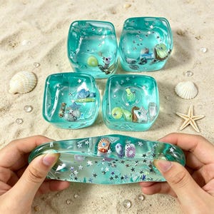 May include: Four translucent teal square resin blocks and one elongated resin piece, all containing embedded miniature seashells and silver star-shaped glitter. The pieces are arranged on a sandy surface with scattered loose seashells.