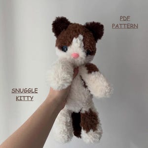 May include: A plush cat toy with white and brown fur, blue eyes, and a pink nose. The text PDF PATTERN and SNUGGLE KITTY is visible. The toy has a soft, fuzzy texture and rounded ears, presented as a digital document for crafting purposes.