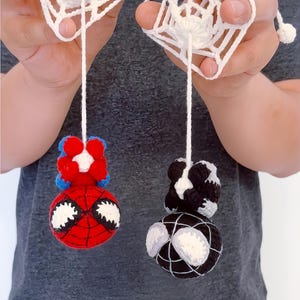 Op de afbeelding: Twee gehaakte garen ornamenten met bolvormige figuren die hangen aan witte gehaakte webachtige vierkanten. Eén figuur is rood en blauw met grote witte ogen, de andere is zwart en wit met bijpassende witte ogen en webdetails.