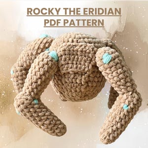 May include: A crocheted spider toy, featuring a tan body and legs with light blue details. The text "ROCKY THE ERIDIAN PDF PATTERN" is displayed above the toy. The toy is made from a soft, textured yarn.