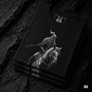 Close up detail of the deep design style graphic print on a soft black fabric fold, showcasing the fine white reflections on the iron helmet, the realistic horse mane texture, and the fluid armor lines. Lifestyle photograph of the Turkish warrior tee stacked neatly within a premium clothing collection to highlight the solid black cotton tone and the high-contrast white fantasy line art. This piece serves as an absolute masterpiece.