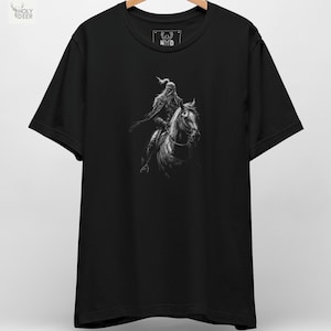 A premium Turkish warrior tee folded neatly on a dark textured rock background, showcasing a highly detailed monochrome illustration of a nomad cavalryman on his horse, centered on the chest. This Attila the Hun shirt captures pure steppe rider apparel energy.