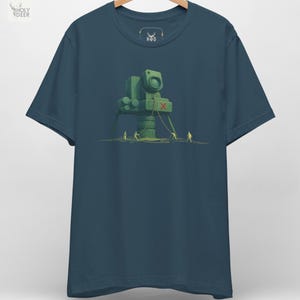 May include: Dark teal t-shirt featuring a green, stylized illustration of a large camera-like structure with three small figures pulling ropes. The design includes a red "X" and is set against a sandy background.