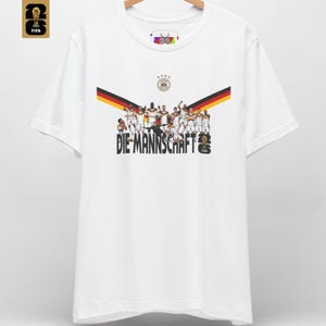 May include: A white short-sleeve t-shirt featuring a graphic of the German national soccer team with the text DIE MANNSCHAFT, a black, red, and gold chevron, the German football crest with four stars, and a FIFA 2026 logo on the chest and corner.