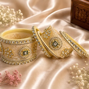 Op de afbeelding: Ivoor en goudkleurige armbanden met ingewikkelde bloemen- en geometrische ontwerpen. De set bevat een brede armband, twee middelgrote armbanden en een dunne armband, allemaal versierd met sprankelende strass-steentjes en een groene vierkante edelsteen.