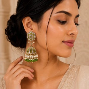 May include: Close-up of ornate green and gold earrings with pearl accents. The earrings feature a floral design at the ear and a bell-shaped drop with multiple layers of green beads and pearls. The earrings are worn by a person.