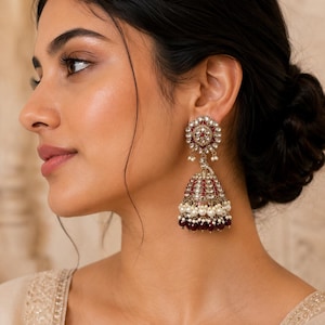 May include: Close-up of a woman wearing ornate, dangling earrings. The earrings feature a floral design with red and clear stones, white pearls, and a deep red bell-shaped bottom. The earrings are a statement piece.