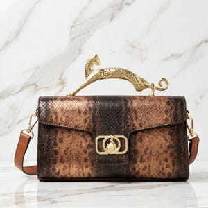 Puede incluir: Un bolso rectangular con estampado de piel de serpiente en tonos marrón y negro, que presenta un asa de metal dorado con forma de pantera saltando. El bolso incluye un cierre rectangular dorado central con un emblema circular y una correa.