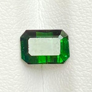May include: A rectangular emerald-cut gemstone with a vibrant, deep green color and faceted surfaces that reflect light, showcasing a clear, transparent center and darker green edges within the stone's geometric structure.