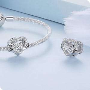 May include: A silver charm bracelet with a heart-shaped charm. The charm has a wing design and is encrusted with small, clear crystals. The word "ver" is visible on the charm. The bracelet is on a light blue background.