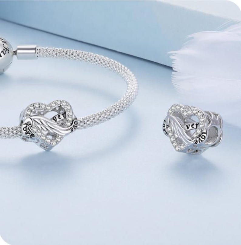 May include: A silver charm bracelet with a heart-shaped charm. The charm has a wing design and is encrusted with small, clear crystals. The word "ver" is visible on the charm. The bracelet is on a light blue background.