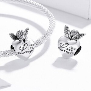 May include: Silver charm beads with an angel design. The charms are heart-shaped and feature the words "Live, Love, Laugh". The angel has detailed wings and a smiling face. The charms are on a silver bracelet.