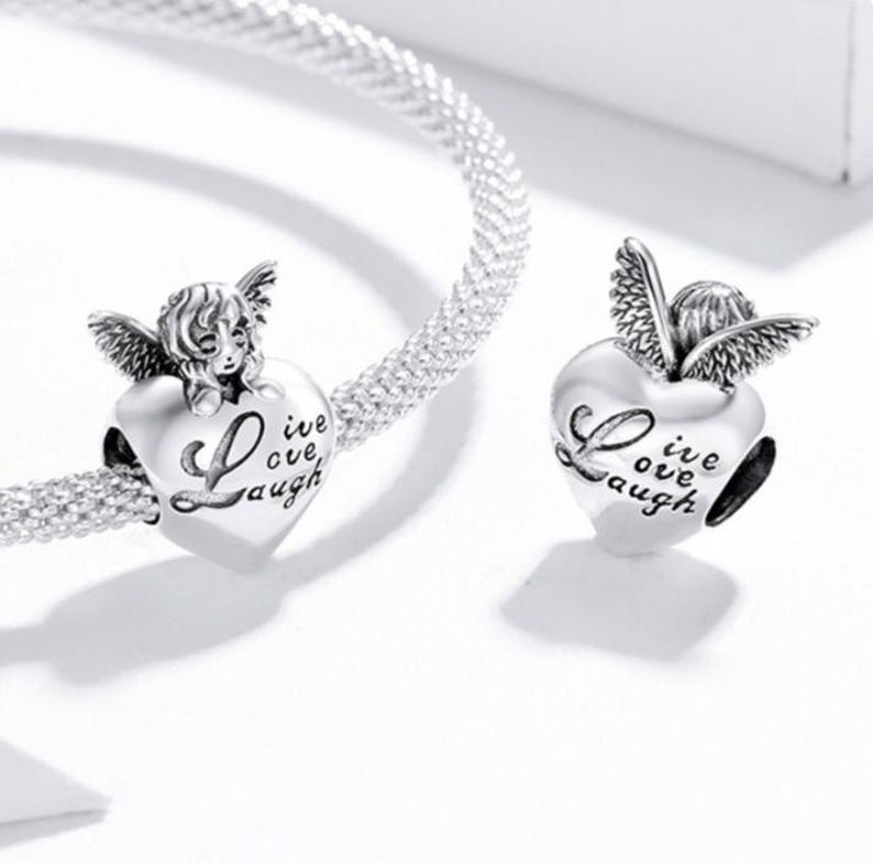 May include: Silver charm beads with an angel design. The charms are heart-shaped and feature the words "Live, Love, Laugh". The angel has detailed wings and a smiling face. The charms are on a silver bracelet.
