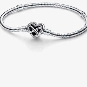 Könnte beinhalten: Ein silbernes Schlangenkettenarmband mit einem herzförmigen Verschluss. Der Verschluss ist mit einem zentralen Unendlichkeitssymbol und kleinen klaren Steinen verziert, die entlang des Herzrandes eingebettet sind. Das Armband hat eine texturierte Oberfläche.