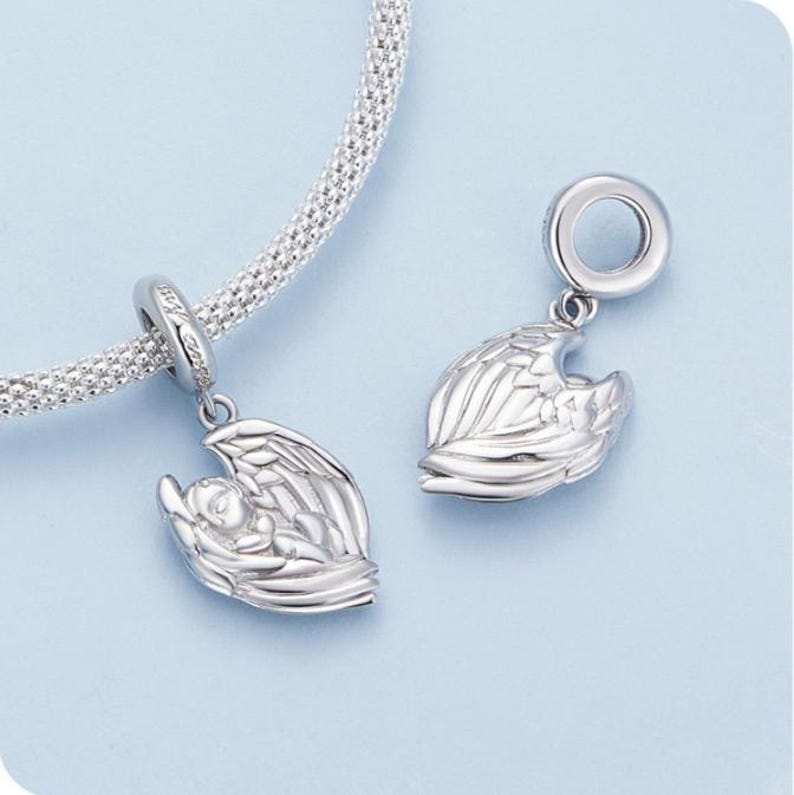 May include: Two silver charms, each featuring a baby cradled in angel wings. The charms are detailed with intricate feather designs and are suspended from a silver chain. The background is a soft blue.