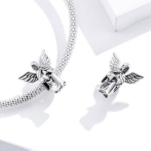 May include: Two silver-colored angel charms with detailed wings. One charm is attached to a textured silver bracelet. The angels are in a flying pose, with intricate detailing on their wings and robes. The background is white.