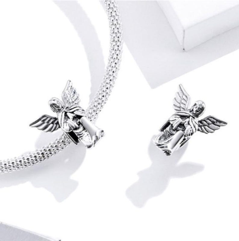May include: Two silver-colored angel charms with detailed wings. One charm is attached to a textured silver bracelet. The angels are in a flying pose, with intricate detailing on their wings and robes. The background is white.