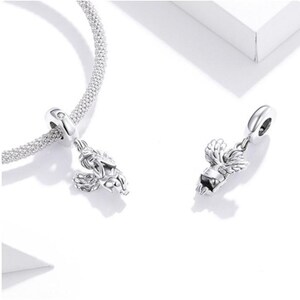 May include: Two silver-colored charm pendants, one attached to a silver chain bracelet. Each charm features a detailed design of a winged figure. The charms are displayed on a white surface, with soft shadows.