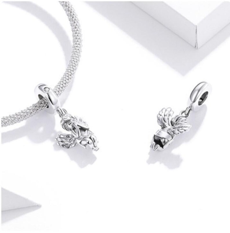 May include: Two silver-colored charm pendants, one attached to a silver chain bracelet. Each charm features a detailed design of a winged figure. The charms are displayed on a white surface, with soft shadows.