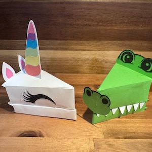 May include: Two papercraft animal-shaped boxes. One is a white unicorn with a rainbow horn and pink ears. The other is a green alligator with white teeth and black eyes. Both are triangular in shape.