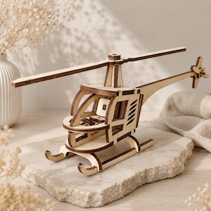 May include: A wooden model helicopter with a natural wood finish. The model features a detailed design, including a main rotor, tail rotor, and cabin. The helicopter is displayed on a light-colored stone surface, with a vase and dried flowers in the background.