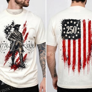 May include: An off-white short-sleeve t-shirt featuring a graphic of a colonial soldier with a rifle over a distressed American flag design. The front displays the years 1776 and 2026, while the back shows a stylized flag with the number 250.
