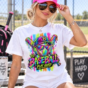 May include: A white short-sleeve t-shirt featuring a colourful graphic of a baseball wearing sunglasses, positioned in front of two crossed baseball bats. The design includes vibrant paint drips and the text BASEBALL PLAY HARD in a bold, multi-coloured font.