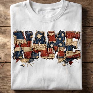 May include: White t-shirt with the word "NAME" in a distressed American flag design. The letters are made of red, white, and blue fabric with a vintage, worn look. The shirt is folded on a wooden surface.