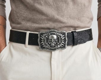 Peut inclure: Une ceinture noire ornée d'un motif floral en relief et d'une boucle rectangulaire en métal argenté. La boucle présente un motif central de crâne entouré de détails en forme de flammes et une bordure avec de petits clous circulaires.