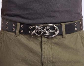 Peut inclure: Ceinture en cuir noir avec une boucle argentée ornée. La ceinture comporte des œillets argentés et est portée avec un pantalon vert olive et un t-shirt gris. La boucle présente un design stylisé unique.