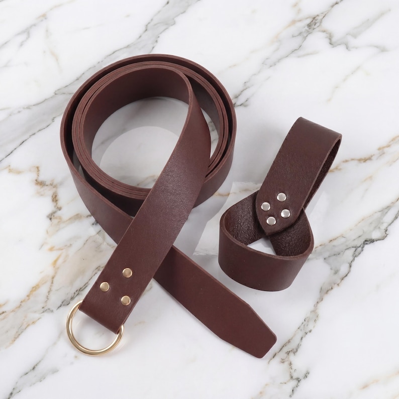 Peut inclure: Une ceinture en cuir marron fonc&eacute; avec une boucle en anneau en m&eacute;tal dor&eacute; et une boucle de ceinture assortie. La ceinture est enroul&eacute;e et la boucle est s&eacute;par&eacute;e, le tout sur une surface en marbre.