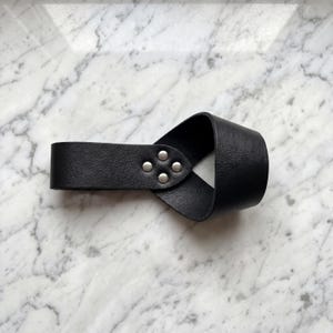 Peut inclure: Manchette de poignet en cuir noir avec un design en boucle. Le bracelet est fix&eacute; avec quatre rivets argent&eacute;s. Le produit est pr&eacute;sent&eacute; sur une surface en marbre blanc et gris.
