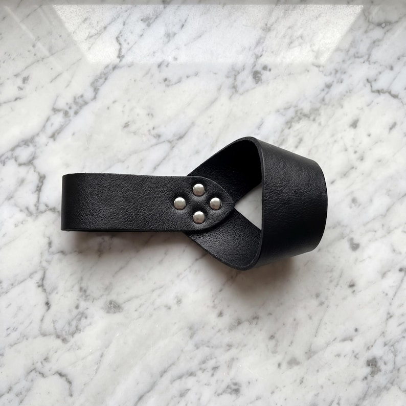 Peut inclure: Manchette de poignet en cuir noir avec un design en boucle. Le bracelet est fix&eacute; avec quatre rivets argent&eacute;s. Le produit est pr&eacute;sent&eacute; sur une surface en marbre blanc et gris.