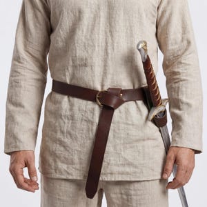 Puede incluir: Un cinturón de cuero marrón con una hebilla dorada y una vaina de espada. La espada tiene una hoja plateada y un mango decorativo. El cinturón se lleva sobre una túnica de lino beige.