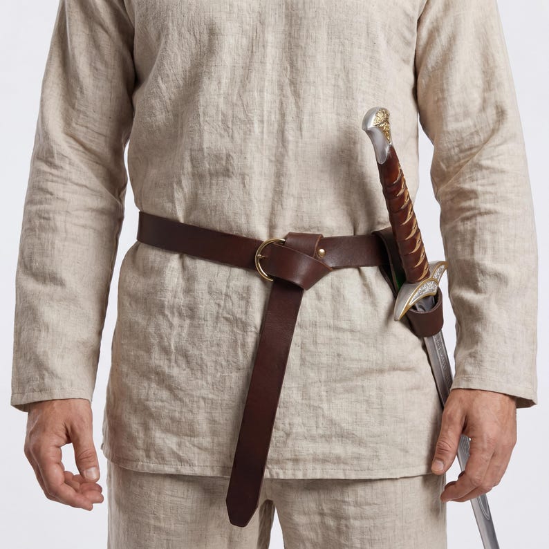 Peut inclure: Une ceinture en cuir marron avec une boucle dor&eacute;e et un fourreau d'&eacute;p&eacute;e. L'&eacute;p&eacute;e a une lame argent&eacute;e et un manche d&eacute;coratif. La ceinture est port&eacute;e sur une tunique en lin beige.