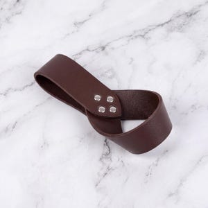 Peut inclure: Une lani&egrave;re en cuir marron fonc&eacute; avec des rivets argent&eacute;s. La lani&egrave;re est boucl&eacute;e et semble &ecirc;tre une poign&eacute;e ou une boucle pour un sac ou un autre accessoire. Le fond est un motif de marbre blanc et gris.