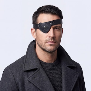 May include: A black leather eye patch with a decorative embossed design and silver-colored rivets. The eye patch is worn over the right eye. The person is wearing a dark gray coat and sweater.