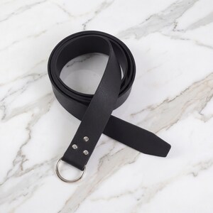 Peut inclure: Une ceinture en cuir noir avec une boucle en anneau en m&eacute;tal argent&eacute;. La ceinture est enroul&eacute;e sur une surface en marbre, mettant en valeur sa texture lisse. La ceinture mesure environ 3,8 cm de large.