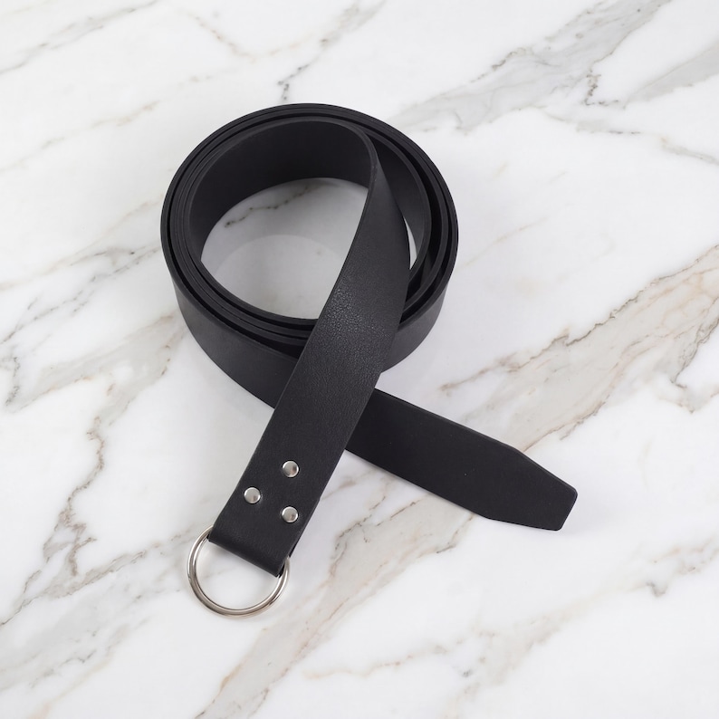Peut inclure: Une ceinture en cuir noir avec une boucle en anneau en m&eacute;tal argent&eacute;. La ceinture est enroul&eacute;e sur une surface en marbre, mettant en valeur sa texture lisse. La ceinture mesure environ 3,8 cm de large.