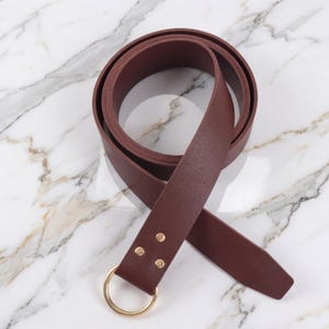Peut inclure: Une ceinture bordeaux avec une boucle circulaire dor&eacute;e et trois petits rivets dor&eacute;s. La ceinture est faite d'un mat&eacute;riau lisse et est pr&eacute;sent&eacute;e sur une surface en marbre blanc et gris. La ceinture est enroul&eacute;e.