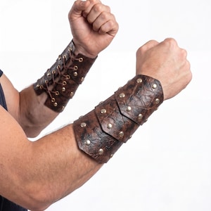 May include: A pair of dark brown textured leather bracers featuring metallic studs and a lace-up closure system. The arm guards are constructed with overlapping panels and reinforced eyelets, providing a structured fit around the forearms.