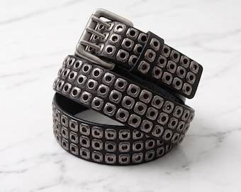 Peut inclure: Une ceinture noire ornée de trois rangées d'œillets métalliques carrés et d'une boucle à double ardillon. La ceinture est enroulée, révélant le motif régulier de ferrures argentées incrustées tout le long de la sangle noire.