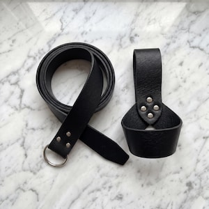 Peut inclure: Une ceinture en cuir noir avec une boucle &agrave; anneau en m&eacute;tal argent&eacute; et des rivets. &Agrave; c&ocirc;t&eacute;, un porte-gobelet en cuir noir avec des rivets argent&eacute;s. Les articles sont pr&eacute;sent&eacute;s sur une surface en marbre blanc et gris.