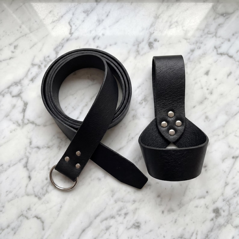 Peut inclure: Une ceinture en cuir noir avec une boucle &agrave; anneau en m&eacute;tal argent&eacute; et des rivets. &Agrave; c&ocirc;t&eacute;, un porte-gobelet en cuir noir avec des rivets argent&eacute;s. Les articles sont pr&eacute;sent&eacute;s sur une surface en marbre blanc et gris.