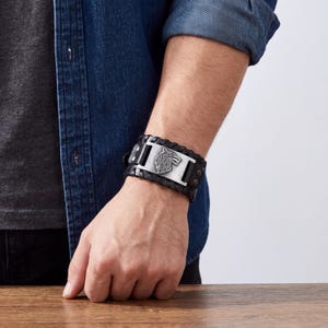 Puede incluir: Pulsera de cuero negro con un emblema de lobo plateado. La pulsera presenta un diseño trenzado y un cierre de hebilla ajustable. El emblema metálico es rectangular y tiene un diseño detallado de cabeza de lobo.
