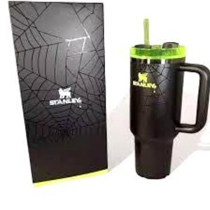 K&ouml;nnte beinhalten: Schwarzer Stanley-Becher mit Griff, gr&uuml;nem Deckel und gr&uuml;nem Strohhalm. Der Becher hat das Stanley-Logo und ein Spinnennetz-Design. Eine passende schwarze Box mit Spinnennetz-Design und dem Stanley-Logo befindet sich neben dem Becher.