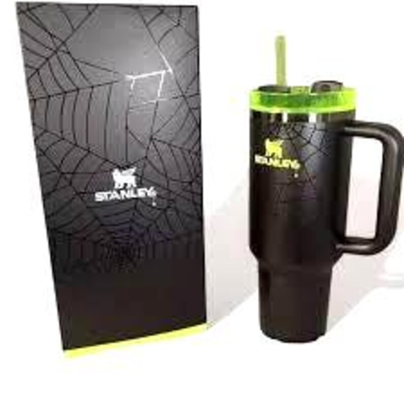 K&ouml;nnte beinhalten: Schwarzer Stanley-Becher mit Griff, gr&uuml;nem Deckel und gr&uuml;nem Strohhalm. Der Becher hat das Stanley-Logo und ein Spinnennetz-Design. Eine passende schwarze Box mit Spinnennetz-Design und dem Stanley-Logo befindet sich neben dem Becher.