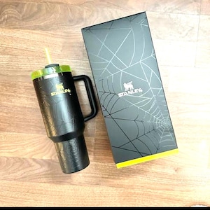 K&ouml;nnte beinhalten: Schwarzer Stanley-Becher mit gr&uuml;nem Deckel und gelbem Strohhalm. Der Becher hat einen Griff und ein Spinnennetz-Design. Die daneben liegende Box hat ebenfalls ein Spinnennetz-Design und das Stanley-Logo.