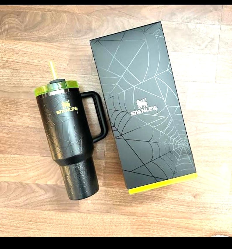 K&ouml;nnte beinhalten: Schwarzer Stanley-Becher mit gr&uuml;nem Deckel und gelbem Strohhalm. Der Becher hat einen Griff und ein Spinnennetz-Design. Die daneben liegende Box hat ebenfalls ein Spinnennetz-Design und das Stanley-Logo.