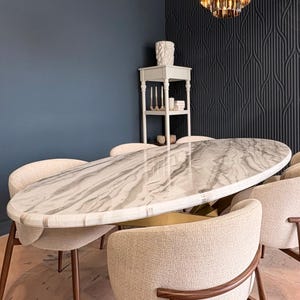 May include: An oval marble dining table with a white and gray pattern, surrounded by beige upholstered chairs with wooden frames. The table is set in a room with dark blue walls and a decorative chandelier.